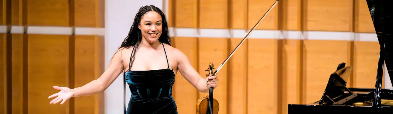 Violinist Ade Williams speaking between pieces at the 2025 Young Performers' Career Advancement Showcase at APAP|NYC 2025. She is wearing a green velvet dress and has dark hair with braids.