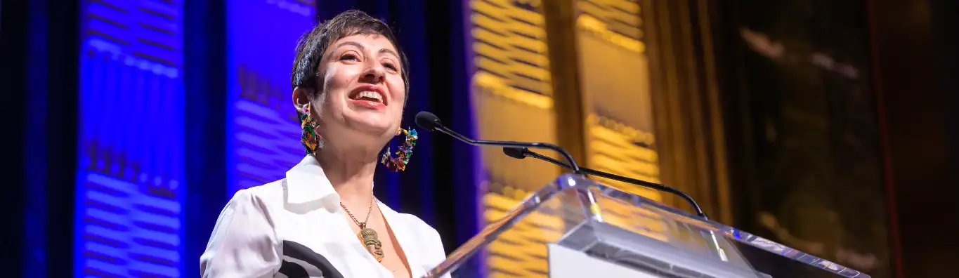 Afa Dworkin speaking at the 2025 APAP Honors Awards Ceremony. She has short brown hair and is wearing a white top and colorful dangly earrings made of fabric.