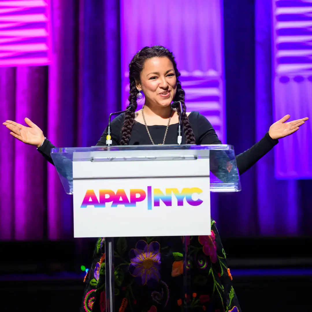 Araceli Poma sharing her story at APAP|NYC 2025. She has dark hair and has her hands outstretched and is wearing a black dress with bright floral emboridery.