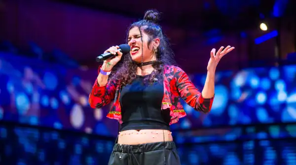 Maruja Limon performing at globalFEST APAP|NYC 2025. Image features a singer with dark curly hair with her left arm up in the air. she is wearing a black outfit with a red cropped sweater.