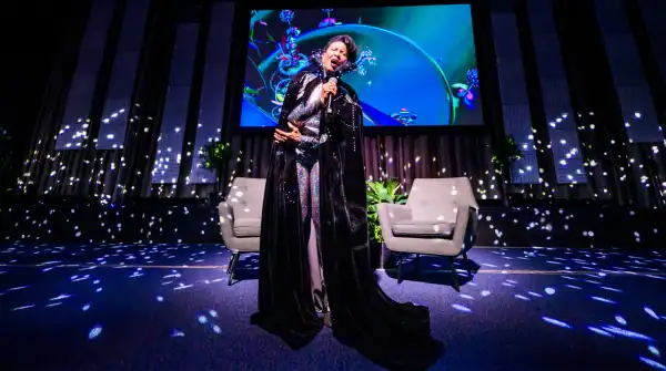 Nona Hendryx performing at APAP|NYC 2025. She is singing while wearing a long black velvet cape with a collar. There are colorful graphics being projected behind her as well as disco lights.