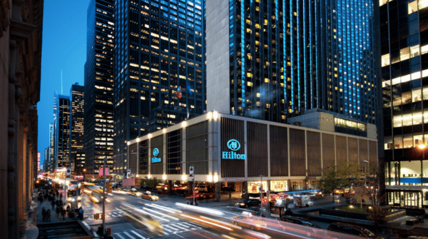 Long-exposure photo of exterior of the New York Hilton Midtown at night with cars streaming down 6th avenue leaving a trail of headlights and breaklights.